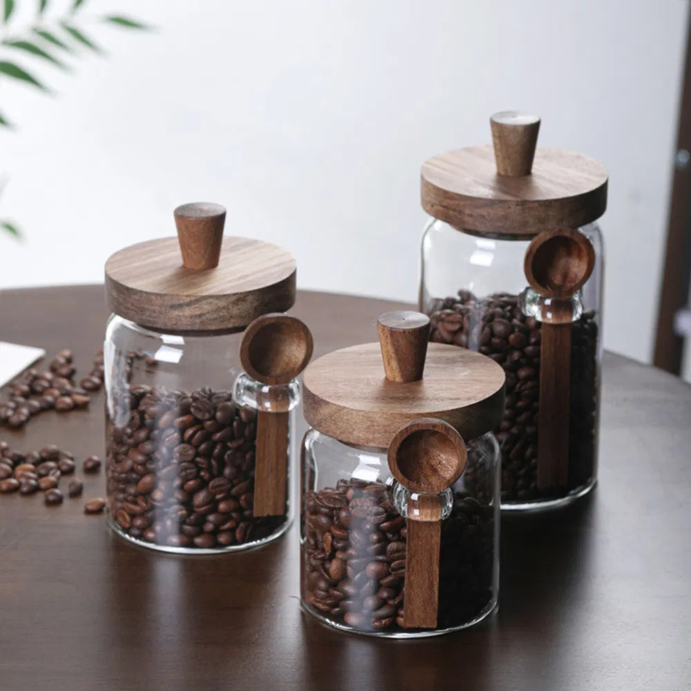 Close-up of three glass jars with coffee beans, wooden lids, and attached wooden spoons on a tabletop