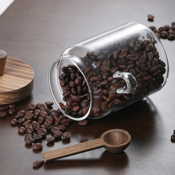 Glass jar tipped on its side spilling coffee beans, with wooden lid and wooden spoon lying beside