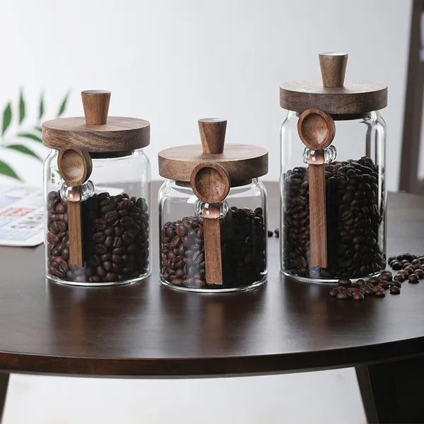 Glass jars with coffee beans and wooden lids displayed on a dark wood table, each with a wooden spoon