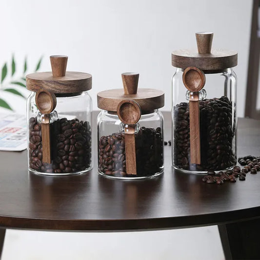 Glass jars with coffee beans and wooden lids displayed on a dark wood table, each with a wooden spoon