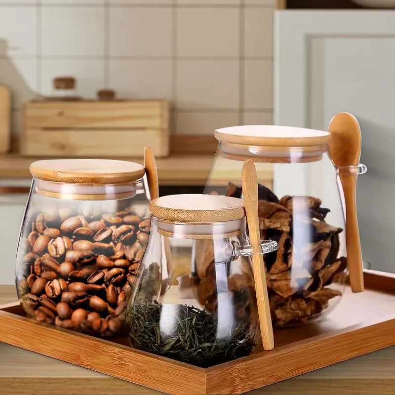 Three glass storage jars with bamboo lids and spoons on a wooden tray filled with coffee beans, dried fruit, and tea