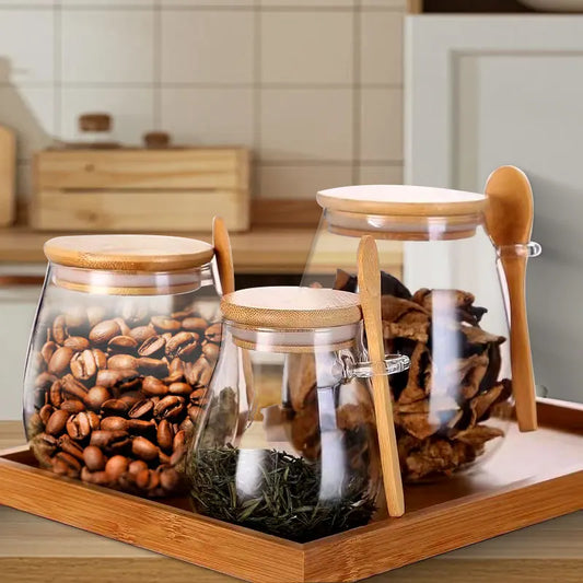 Three glass storage jars with bamboo lids and spoons on a wooden tray filled with coffee beans, dried fruit, and tea