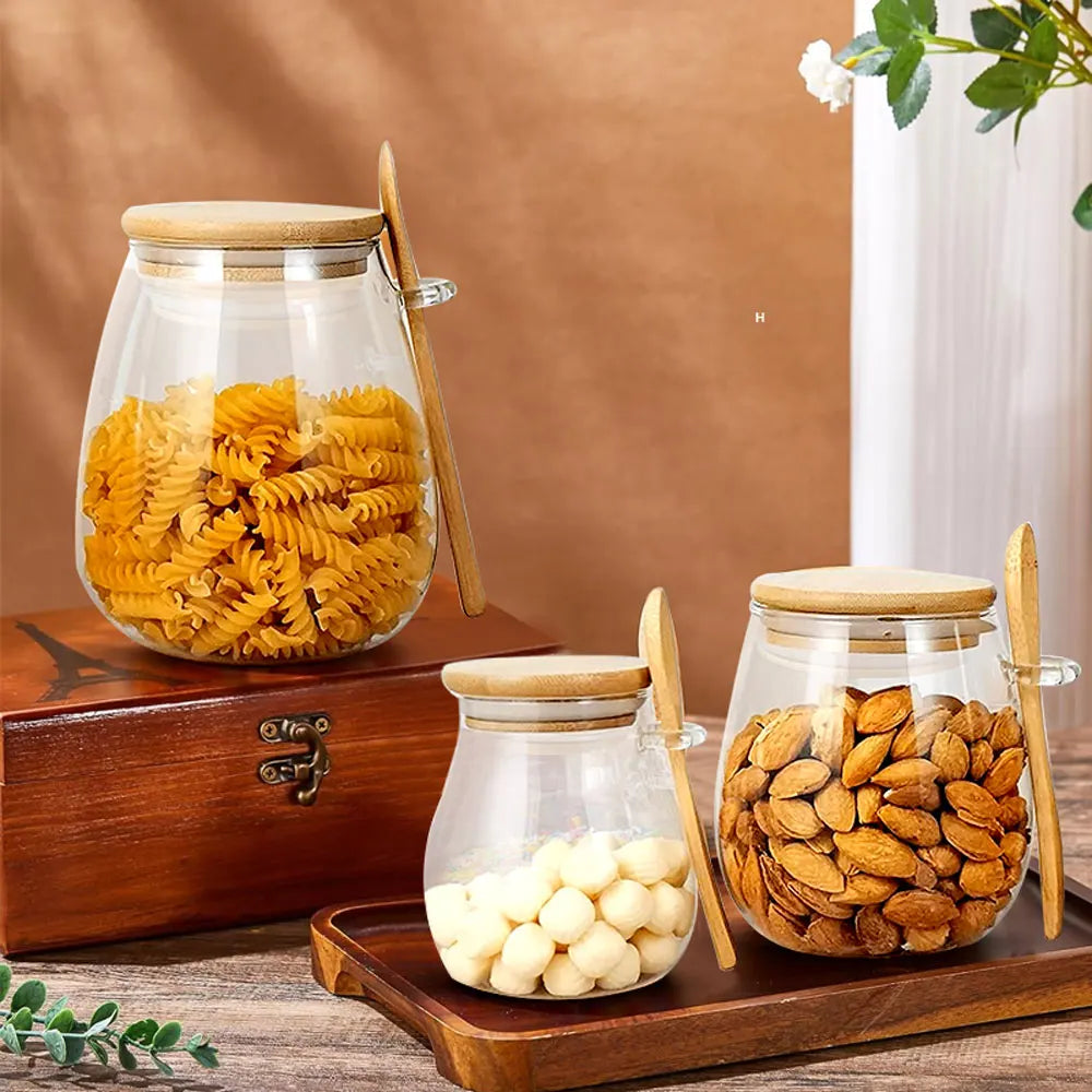 Three glass storage jars with bamboo lids and spoons on a wooden surface, filled with pasta, nuts, and treats